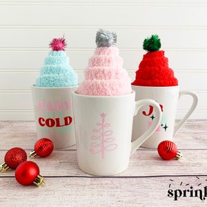 May include: Three white mugs with different designs. The mugs are filled with fluffy socks that look like winter hats. The socks are pink, blue, and red. The mugs are sitting on a wooden surface with red Christmas ornaments.