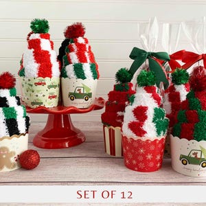 May include: A set of 12 cupcake-shaped socks featuring red, green, white, and black fuzzy textures topped with pom-poms. The socks are presented in paper liners decorated with gingerbread men, trucks carrying trees, and snowflakes, with some packaged in clear bags.