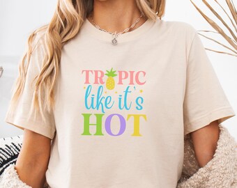 May include: Beige t-shirt with the text "Tropic like it's HOT" in colorful letters and a pineapple graphic. The model is wearing denim shorts and a beige sweater. The shirt is a casual, summery style.