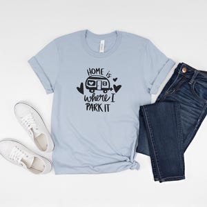May include: A light blue t-shirt with the text "HOME is where I PARK IT" in black, along with a graphic of a camper and hearts. The shirt is paired with dark blue jeans and white sneakers.