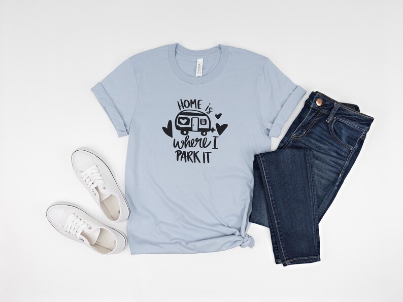 May include: A light blue t-shirt with the text "HOME is where I PARK IT" in black, along with a graphic of a camper and hearts. The shirt is paired with dark blue jeans and white sneakers.
