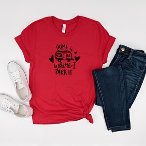 May include: A red t-shirt with the phrase "Home is where I park it" in black, along with a graphic of a camper and hearts. The shirt is paired with dark blue jeans and white sneakers.