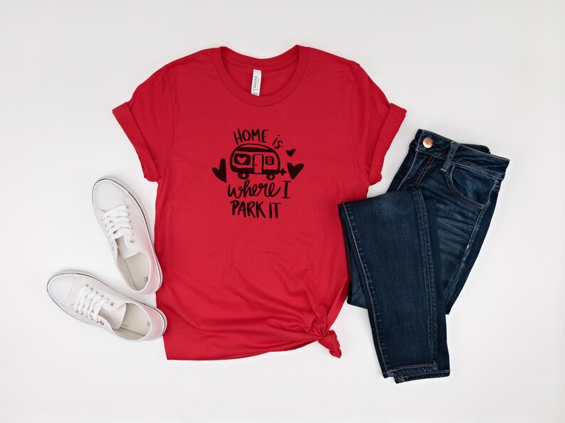 May include: A red t-shirt with the phrase "Home is where I park it" in black, along with a graphic of a camper and hearts. The shirt is paired with dark blue jeans and white sneakers.
