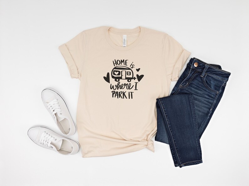 May include: A beige t-shirt with the text "Home is where I park it" and a camper graphic, paired with dark blue jeans and white sneakers. The shirt is folded at the bottom. The items are arranged on a white surface.