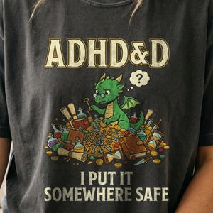 May include: A charcoal gray t-shirt featuring a graphic of a green dragon wearing glasses, sitting on a pile of gold coins, keys, books, and potions. The text reads ADHD&D at the top and I PUT IT SOMEWHERE SAFE at the bottom in a light font.