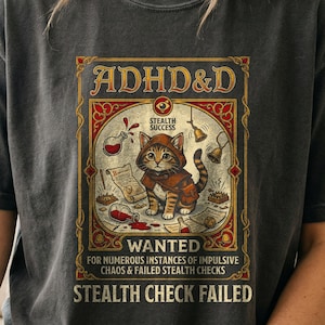 May include: A charcoal gray t-shirt featuring a graphic print of a tabby cat wearing a hooded cloak. The text reads ADHD&D, STEALTH SUCCESS, WANTED FOR NUMEROUS INSTANCES OF IMPULSIVE CHAOS & FAILED STEALTH CHECKS, and STEALTH CHECK FAILED.