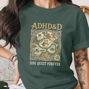 Puede incluir: Una camiseta de manga corta de color verde oscuro con un estampado gr&aacute;fico que incluye el texto ADHD&D y SIDE QUEST FOREVER. El dise&ntilde;o muestra un gato central sosteniendo una espada, rodeado por un mapa circular con caminos, una taberna y un drag&oacute;n.