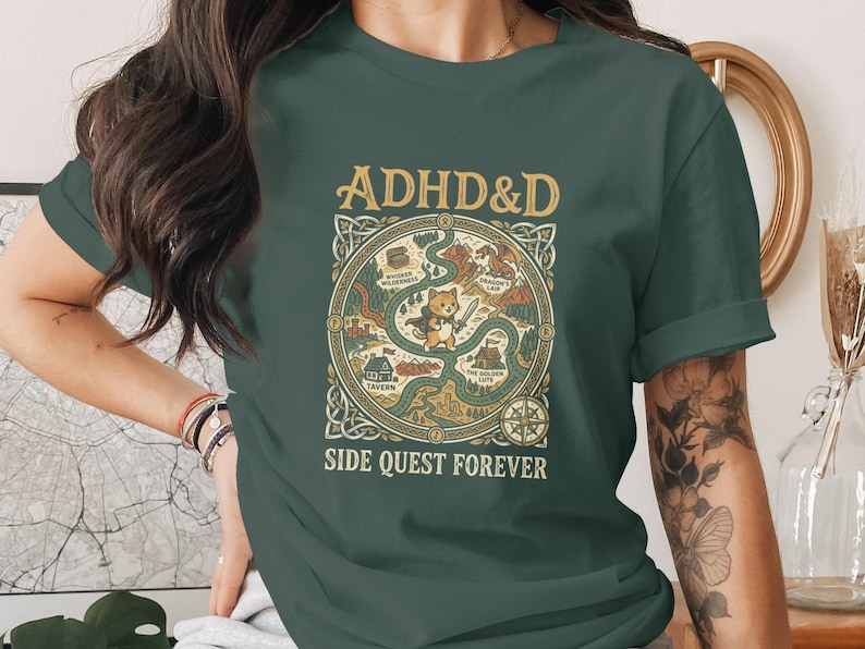 Puede incluir: Una camiseta de manga corta de color verde oscuro con un estampado gr&aacute;fico que incluye el texto ADHD&D y SIDE QUEST FOREVER. El dise&ntilde;o muestra un gato central sosteniendo una espada, rodeado por un mapa circular con caminos, una taberna y un drag&oacute;n.