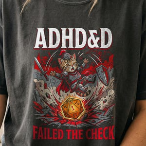 May include: A charcoal gray t-shirt featuring a graphic of a tabby cat dressed in knight armor, holding a sword and shield, leaping over a twenty-sided die showing a one. The text reads ADHD&D at the top and FAILED THE CHECK at the bottom.