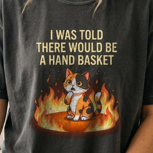 以下が含まれることがあります： チャコールグレーのTシャツで、オレンジと黄色の炎の輪の中に座っている三毛猫のグラフィックが描かれています。猫の上には、薄い黄色のサンセリフフォントで「I WAS TOLD THERE WOULD BE A HAND BASKET」というテキストがプリントされています。