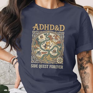 Puede incluir: Una camiseta azul marino de manga corta con un estampado gr&aacute;fico de un mapa de fantas&iacute;a con un gato central que sostiene una espada. El texto dice ADHD&D en la parte superior y SIDE QUEST FOREVER en la inferior, con un borde celta circular.