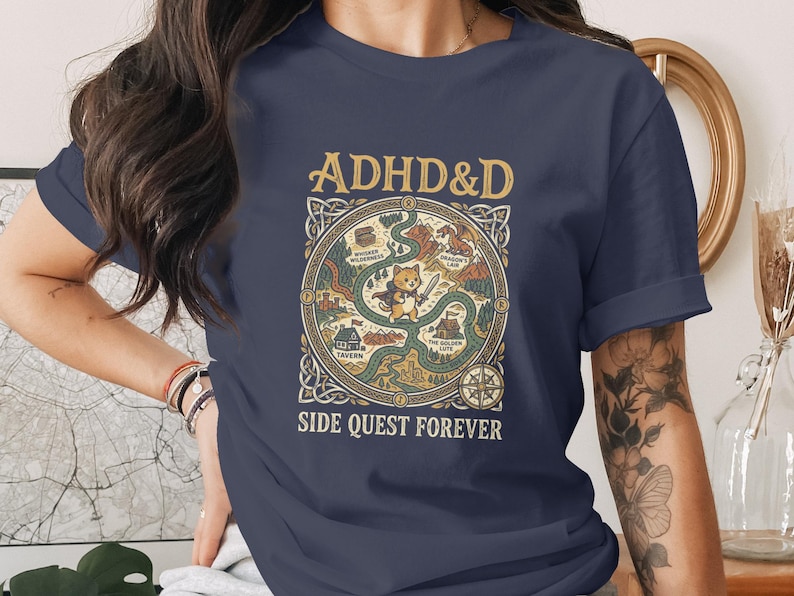 Puede incluir: Una camiseta azul marino de manga corta con un estampado gr&aacute;fico de un mapa de fantas&iacute;a con un gato central que sostiene una espada. El texto dice ADHD&D en la parte superior y SIDE QUEST FOREVER en la inferior, con un borde celta circular.