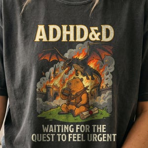 May include: A charcoal gray t-shirt featuring a graphic print with the text ADHD&D at the top and WAITING FOR THE QUEST TO FEEL URGENT at the bottom. The image shows a capybara holding a mug in front of a burning castle with a dragon flying above.