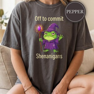 Peut inclure: Un t-shirt gris anthracite avec une illustration de grenouille verte déguisée en magicien, portant une robe et un chapeau violets, tenant un bâton avec un cristal violet brillant. Le texte indique Off to commit Shenanigans. Une étiquette PEPPER.