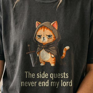 May include: A charcoal gray t-shirt featuring a graphic of an orange tabby cat wearing a dark hooded cloak and holding a sword. Below the cat, the text reads, The side quests never end my lord. The design has a distressed, vintage appearance.