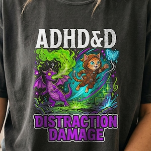 May include: A charcoal gray t-shirt featuring a graphic print with the text ADHD&D DISTRACTION DAMAGE. The design shows an orange cat in a hooded cloak leaping toward a purple dragon breathing green smoke, surrounded by blue magical effects.