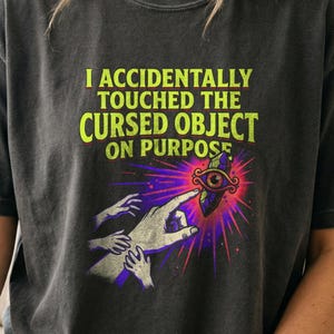 May include: A charcoal gray t-shirt featuring the lime green text I ACCIDENTALLY TOUCHED THE CURSED OBJECT ON PURPOSE. Below the text is a graphic of a hand reaching toward a floating purple crystal with a single eye, surrounded by pink rays.