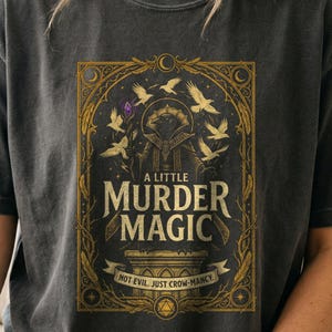 Könnte beinhalten: Ein anthrazitfarbenes T-Shirt mit einer goldfarbenen Grafik eines Raben in einer Robe, der einen Stab mit einem violetten Kristall hält. Der Text lautet A LITTLE MURDER MAGIC und NOT EVIL. JUST CROW-MANCY. Das Design ist von fliegenden Raben und verzierten Rändern umgeben.