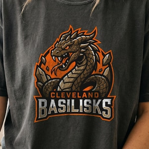 May include: A charcoal gray t-shirt featuring a graphic of a brown basilisk with orange eyes and a forked tongue. The design includes sharp rock elements and the text CLEVELAND BASILISKS in bold, blocky lettering set against an orange circular backdrop.