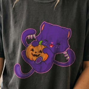 May include: A charcoal gray t-shirt featuring a graphic illustration of a purple cat with sharp claws holding an orange jack-o'-lantern pumpkin. The cat has a smiling expression with visible fangs, and the design is outlined in a bright pink color.