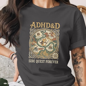 Puede incluir: Una camiseta gris oscuro con un gr&aacute;fico de mapa de fantas&iacute;a circular con el texto ADHD&D en la parte superior y SIDE QUEST FOREVER en la parte inferior. El centro muestra un peque&ntilde;o personaje de gato con una espada, rodeado de lugares.