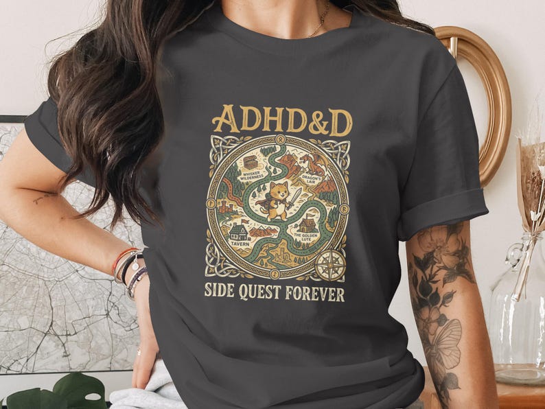 Puede incluir: Una camiseta gris oscuro con un gr&aacute;fico de mapa de fantas&iacute;a circular con el texto ADHD&D en la parte superior y SIDE QUEST FOREVER en la parte inferior. El centro muestra un peque&ntilde;o personaje de gato con una espada, rodeado de lugares.