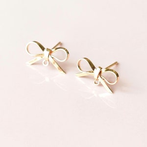 May include: A pair of gold-coloured bow-shaped stud earrings. The earrings are made of a shiny metal and have a delicate, feminine design. The bows are detailed with loops and a knot in the centre. The earrings are on a light pink background.