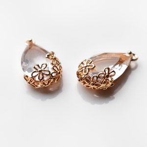 May include: Two teardrop-shaped pendants with a clear, faceted center and a gold-colored floral design around the top. The pendants are set against a white background, showcasing their delicate details.