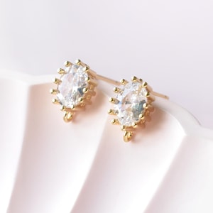 May include: A pair of gold-colored stud earrings. Each earring features an oval-shaped, clear gemstone surrounded by a gold-colored frame with small, pointed accents. The earrings have a post and loop design.