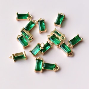 May include: A group of green emerald-shaped charms with gold-toned metal settings and jump rings. The charms are scattered on a white surface.