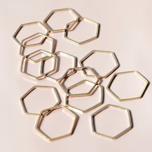 May include: A group of gold-toned hexagonal rings interlocked together. The rings are arranged in a random pattern and are scattered on a light pink background.