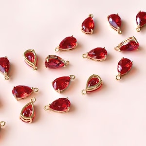 May include: A collection of red teardrop-shaped crystal charms with gold-toned metal loops. The charms are scattered on a light pink surface.