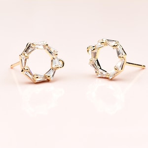 May include: A pair of gold-colored stud earrings. Each earring features a geometric design with a circular shape, adorned with clear, rectangular-cut crystals. The earrings are set against a soft pink background.