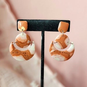 May include: A pair of dangle earrings with a peach, white and gold marbled design. The earrings feature a crescent moon shape with a smaller circle on top.