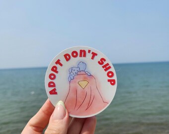 以下が含まれることがあります： 赤い文字で「ADOPT DON'T SHOP」と書かれた丸いステッカー。犬を抱きかかえるマント姿の人物のイラスト。背景にはビーチと海が描かれています。