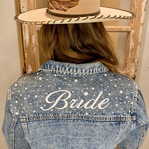 May include: A light blue denim jacket with pearl embellishments and the word "Bride" embroidered on the back.