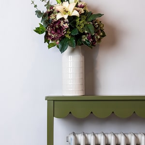 May include: A white ceramic vase with a textured surface holds a bouquet of white lilies and purple hydrangeas. The vase sits on a green table with a scalloped edge. A white radiator is visible behind the table.