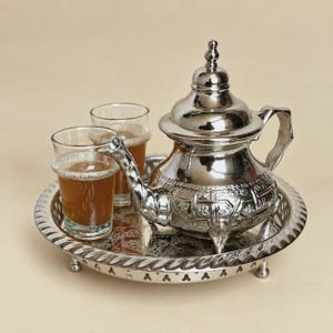 May include: A polished silver-tone metal teapot with an ornate embossed pattern, a pointed lid, and a curved handle. It sits on a matching round tray with a scalloped edge alongside two clear glass tumblers filled with a brown tea beverage.