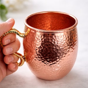 May include: A copper Moscow Mule mug with a hammered texture and a gold-colored handle. The mug has a rounded body and a wide opening. The handle is ornate and curved, providing a comfortable grip. The copper color gives the mug a classic and elegant look.