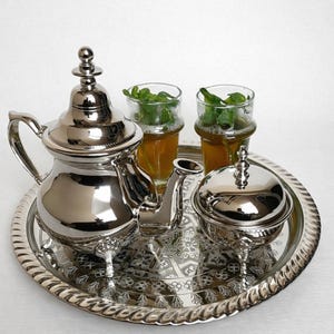 Pode incluir: Um conjunto de chá prateado com uma chaleira, açucareiro e dois copos de chá com folhas de hortelã, apresentado numa bandeja decorativa. O conjunto tem detalhes ornamentados e uma superfície refletora, criando um visual clássico e elegante.
