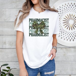 May include: A white short-sleeved t-shirt featuring the word PHARMACIST repeated in dark green text. Centered over the text is a large, detailed bow with a green and brown camouflage pattern. The design is printed on the front of the shirt.