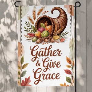 Könnte beinhalten: Eine Gartenflagge mit einer Aquarellillustration eines Füllhorns, das mit Kürbissen, Kürbissen und Eicheln überquillt. Die Flagge trägt in brauner Schrift die Aufschrift "Gather & Give Grace". Herbstblätter und Weizenähren umrahmen das Design.