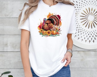 Pode incluir: Camiseta branca com uma ilustração colorida de um peru rodeado por abóboras, girassóis e folhagens de outono. O peru tem penas marrons, laranjas e vermelhas. Um design festivo para o Dia de Ação de Graças.