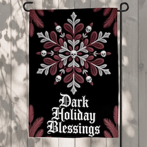 Könnte beinhalten: Eine schwarze Gartenflagge mit einem zentralen schneeflockenartigen Design aus weißen und dunkelroten Blättern, durchsetzt mit kleinen weißen Totenköpfen. Der Text "Dark Holiday Blessings" ist unten in einer weißen gotischen Schrift gedruckt.