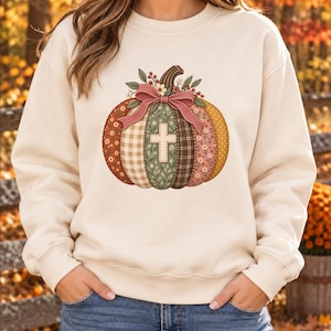 Könnte beinhalten: Ein cremefarbenes Sweatshirt mit Rundhalsausschnitt und einem zentralen Kürbis-Motiv, das aus verschiedenen gemusterten Stoffsegmenten in Orange-, Braun- und Grüntönen besteht, verziert mit einer rosa Schleife, Blumen und einem weißen Kreuz.