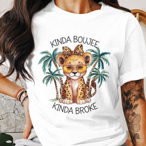 May include: White t-shirt featuring a cartoon cheetah wearing sunglasses and a bow, with the text "Kinda Boujee Kinda Broke". Palm trees are on either side of the cheetah. The shirt is a casual wear item.