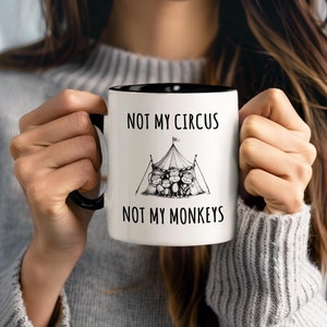 Puede incluir: Taza de cerámica blanca con borde y asa negros. La taza tiene una ilustración en blanco y negro de una carpa de circo con monos dentro. El texto "Not my circus, not my monkeys" está impreso en la taza.