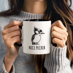 Könnte beinhalten: Eine weiße Kaffeetasse mit schwarzem Rand und Henkel. Die Tasse zeigt eine Schwarzweißillustration eines Spechts mit dem Text "Nice Pecker" darunter.