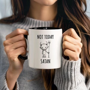 Può includere: Tazza in ceramica bianca con interno e manico neri. La tazza presenta la scritta "NOT TODAY SATAN" sopra un'illustrazione di un diavolo dei cartoni animati che tiene un forcone. La tazza è tenuta da una persona che indossa un maglione grigio.