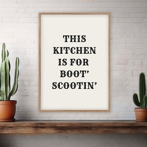 May include: A framed print with the text "This Kitchen is for Boot' Scootin'" hanging on a white brick wall. Two green cactus plants are on a wooden shelf below the print.
